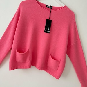 May include: A vibrant pink sweater with a crew neck and two front pockets. The long-sleeved jumper has a brand tag attached. It is displayed on a white hanger against a white backdrop.