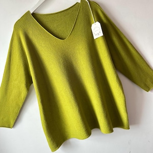 May include: A lime green V-neck jumper with long sleeves, displayed on a white hanger. The knit garment features a relaxed fit and a small white rectangular paper tag attached to the neckline with a thin string, showing faint printed text.