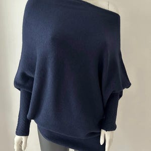 May include: A navy blue, off-the-shoulder jumper with long sleeves. The jumper has a loose, draped design with a wide neckline and textured sleeves. The garment is displayed on a mannequin.