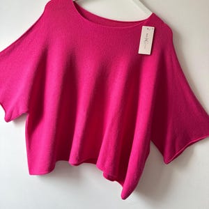 May include: A bright fuchsia knit top with a wide, loose fit, short sleeves, and a rounded neckline. The garment features a rolled hem detail and is displayed on a white hanger with a rectangular tag attached that reads NEW COLLECTION.