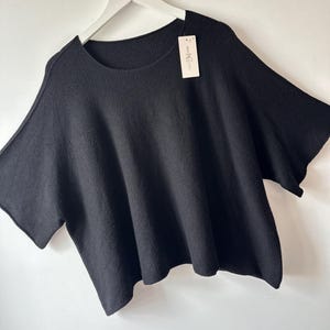 May include: A black short-sleeved jumper with a wide, boxy fit and a round neckline. The garment features a textured knit fabric and includes a small white rectangular brand tag attached to the collar area, hanging from the neckline.