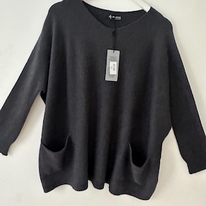 May include: A dark grey sweater with a V-neck and two front pockets. The long-sleeved top has a relaxed fit and a tag attached near the neckline. The sweater is hanging on a white hanger.
