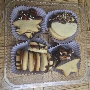 May include: A clear plastic container holding four assorted cookies. Two star-shaped cookies are topped with chocolate and sprinkles. Two round cookies are half-dipped in chocolate and nuts. The other cookies are drizzled with chocolate.