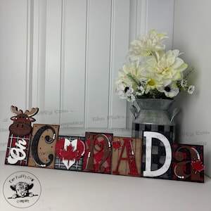 May include: Wooden sign spelling "CANADA" in block letters with a red heart with a maple leaf in the centre. The letters are decorated with red, white and black plaid, polka dots, and glitter. A moose figurine is on the left side of the sign.