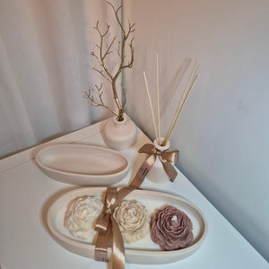 May include: A display of home decor items. It features a small vase with a gold branch, an oval tray, a reed diffuser, and a tray with three flower-shaped candles in cream, beige, and mauve. The items are arranged on a white surface.