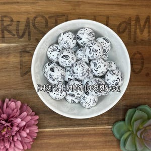 May include: A white ceramic bowl filled with white beads featuring a black paisley design. The beads are scattered inside the bowl. The text "Rusty Llama Design Co." is printed on the bowl. The bowl is on a wooden surface.