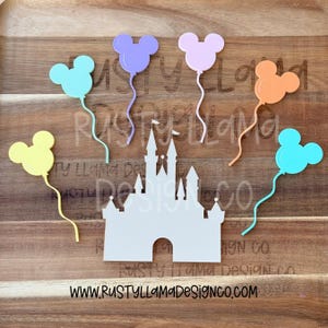May include: A wooden tray displays a white castle cutout and six colorful Mickey Mouse-shaped balloon cutouts in yellow, blue, purple, pink, and orange. The website address www.rustyllamadesignco.com is printed at the bottom.