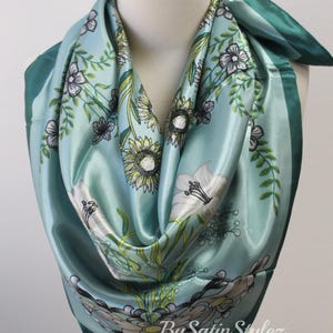 May include: A teal blue satin scarf with a floral print featuring white, yellow, and green flowers. The scarf has a dark teal border and the text "By Satin Stylez" is printed in the bottom right corner.