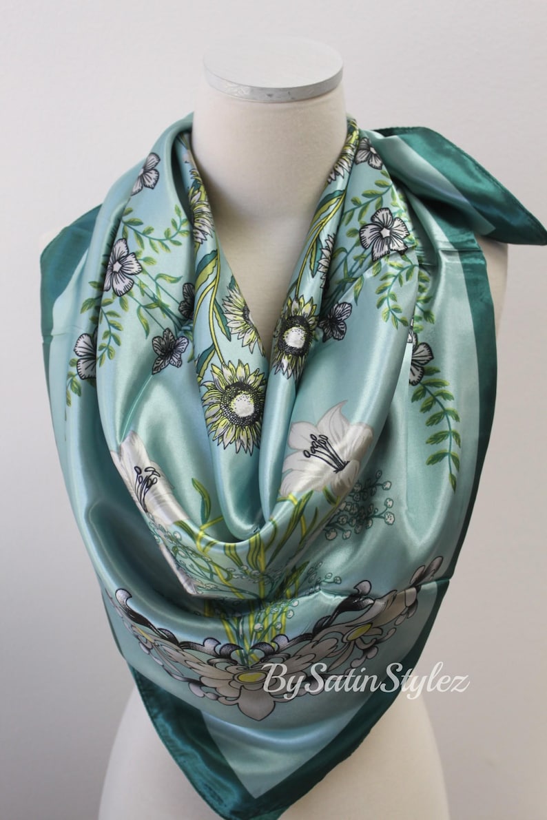May include: A teal blue satin scarf with a floral print featuring white, yellow, and green flowers. The scarf has a dark teal border and the text "By Satin Stylez" is printed in the bottom right corner.