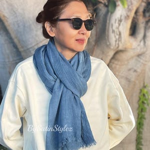 May include: A woman wearing a white sweater and a light blue scarf with a frayed edge. She is wearing sunglasses and looking to the right.