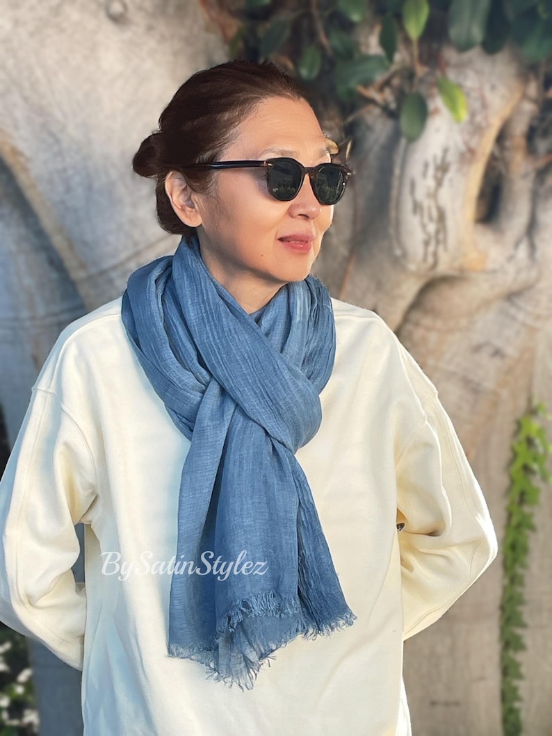 May include: A woman wearing a white sweater and a light blue scarf with a frayed edge. She is wearing sunglasses and looking to the right.