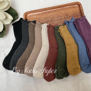 May include: A collection of ten pairs of socks in various colors, including black, gray, brown, cream, red, green, yellow, blue, and purple. The socks are arranged on a wooden tray. The text "By Satin Stylerz" is visible.