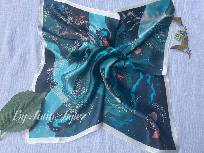 May include: Square silk scarf with an ocean-themed design. The scarf features a dark teal background with illustrations of coral, starfish, and other marine life in shades of teal, blue, and orange. The scarf has a white border and the text "By Satin Stylez".
