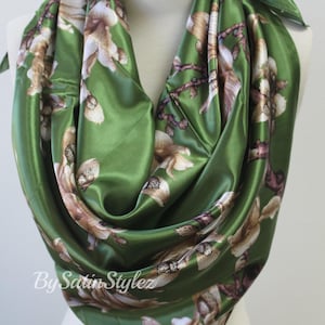 May include: A green satin scarf with a floral print of white and brown flowers. The scarf is folded and draped over a mannequin's neck. The text "By Satin Stylez" is visible in the bottom left corner of the image.