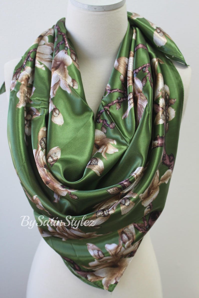 May include: A green satin scarf with a floral print of white and brown flowers. The scarf is folded and draped over a mannequin's neck. The text "By Satin Stylez" is visible in the bottom left corner of the image.