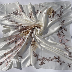 May include: A white satin scarf with a floral print of brown and pink flowers and leaves. The scarf is folded and draped on a white surface.