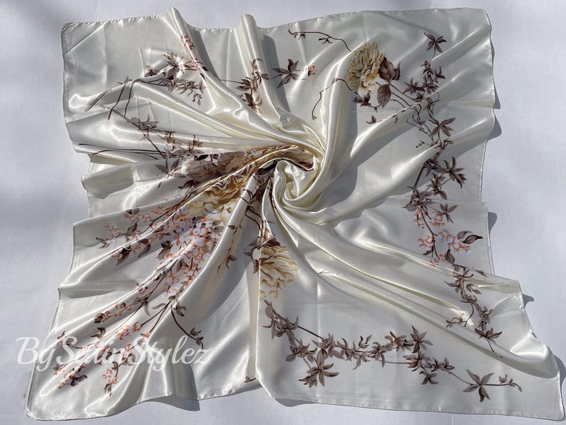 May include: A white satin scarf with a floral print of brown and pink flowers and leaves. The scarf is folded and draped on a white surface.