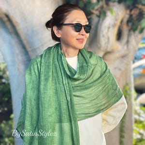 May include: A green, textured scarf with a subtle ombre effect. The scarf is draped around a person's neck and shoulders. The person is wearing a white shirt and sunglasses.