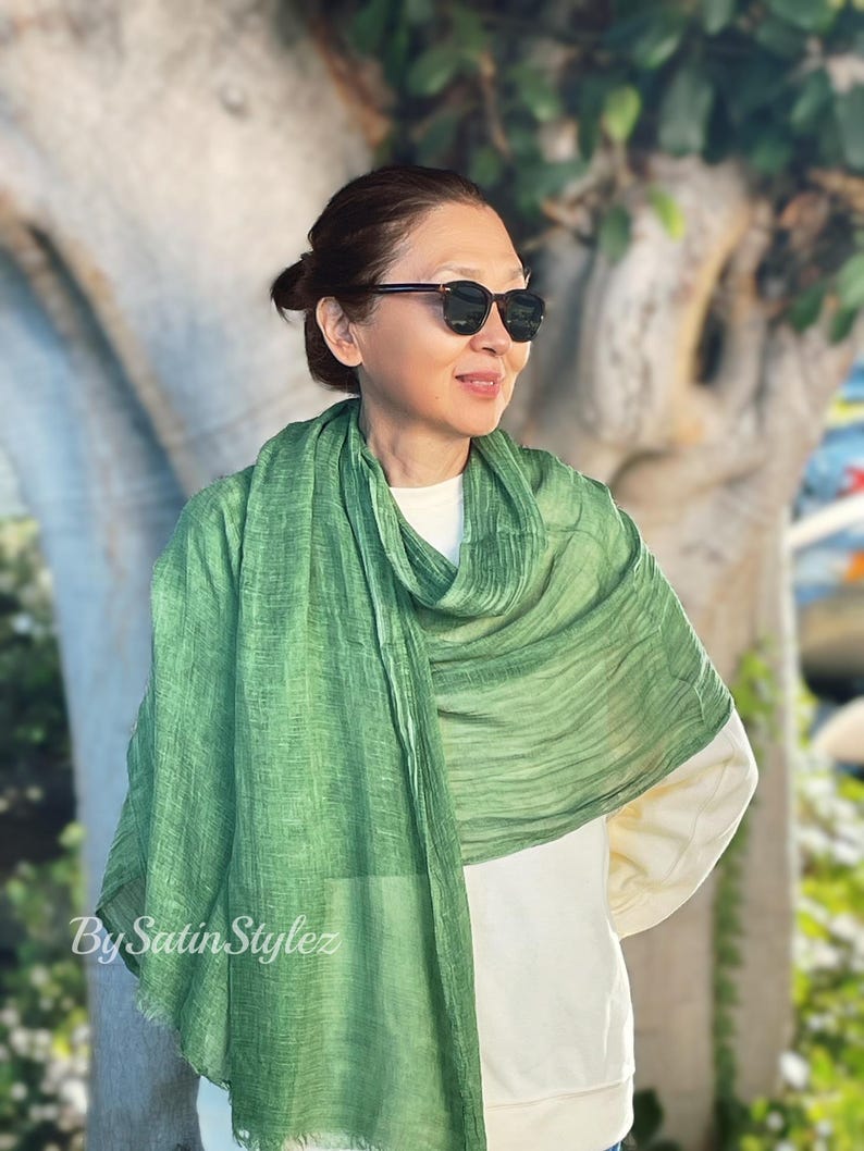 May include: A green, textured scarf with a subtle ombre effect. The scarf is draped around a person's neck and shoulders. The person is wearing a white shirt and sunglasses.