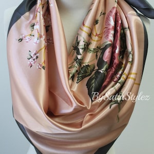 May include: A pink satin scarf with a floral print and black trim. The scarf is folded and displayed on a mannequin.