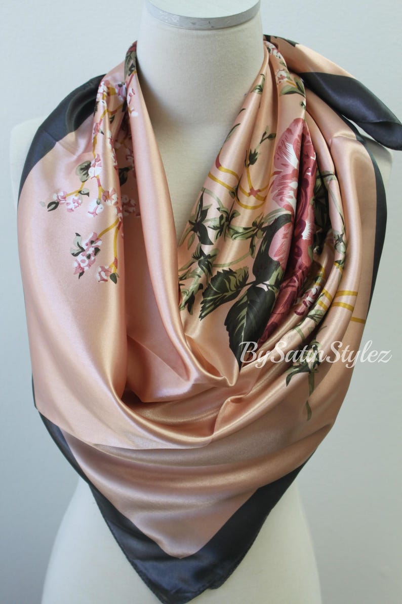 May include: A pink satin scarf with a floral print and black trim. The scarf is folded and displayed on a mannequin.