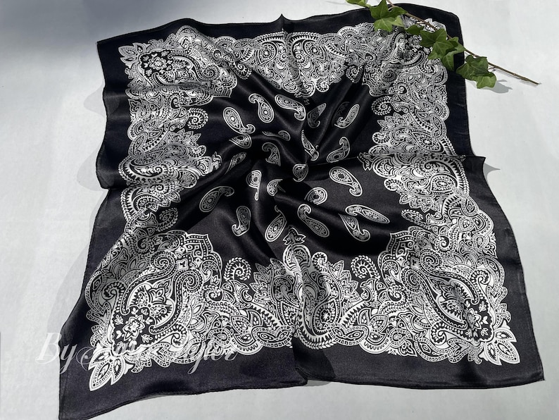 May include: Black satin bandana with a white paisley pattern. The bandana is folded and laid flat on a white surface.