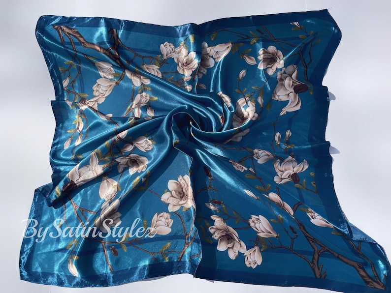 May include: A teal blue satin scarf with a white floral print. The scarf has a white border and the text "By Satin Stylez" is printed on the bottom left corner.