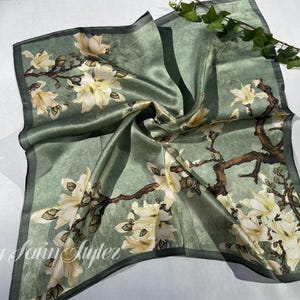 May include: A square, sage green silk scarf with a floral design. The scarf features white and cream-colored flowers with brown branches. The edges are trimmed with a darker green border. The text "By Satin Styler" is visible.