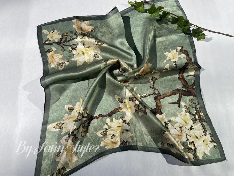 May include: A square, sage green silk scarf with a floral design. The scarf features white and cream-colored flowers with brown branches. The edges are trimmed with a darker green border. The text "By Satin Styler" is visible.