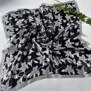 May include: Black and white floral silk scarf. The scarf features a repeating pattern of white flowers and leaves on a black background. The edges are trimmed with a silver border. A sprig of greenery rests on the scarf.