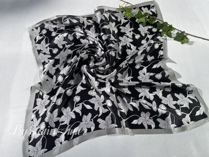 May include: Black and white floral silk scarf. The scarf features a repeating pattern of white flowers and leaves on a black background. The edges are trimmed with a silver border. A sprig of greenery rests on the scarf.