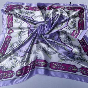 May include: A purple satin scarf with a white background and a floral pattern. The pattern features small white flowers with green leaves and a pink border.