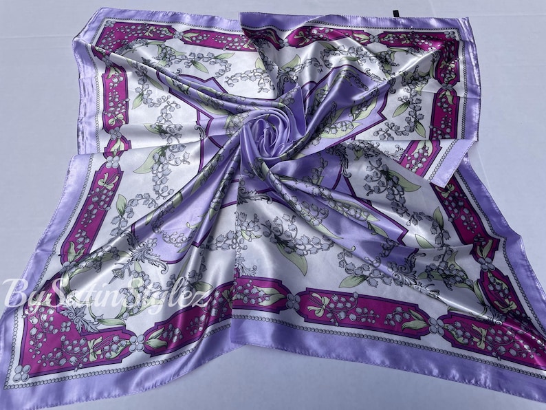 May include: A purple satin scarf with a white background and a floral pattern. The pattern features small white flowers with green leaves and a pink border.