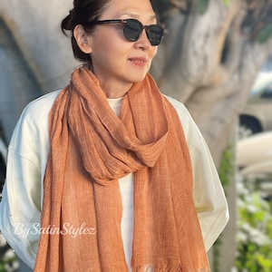 May include: A rust-coloured, fringed scarf made of a soft, textured fabric. The scarf is draped around the neck of a person wearing a white shirt and sunglasses.