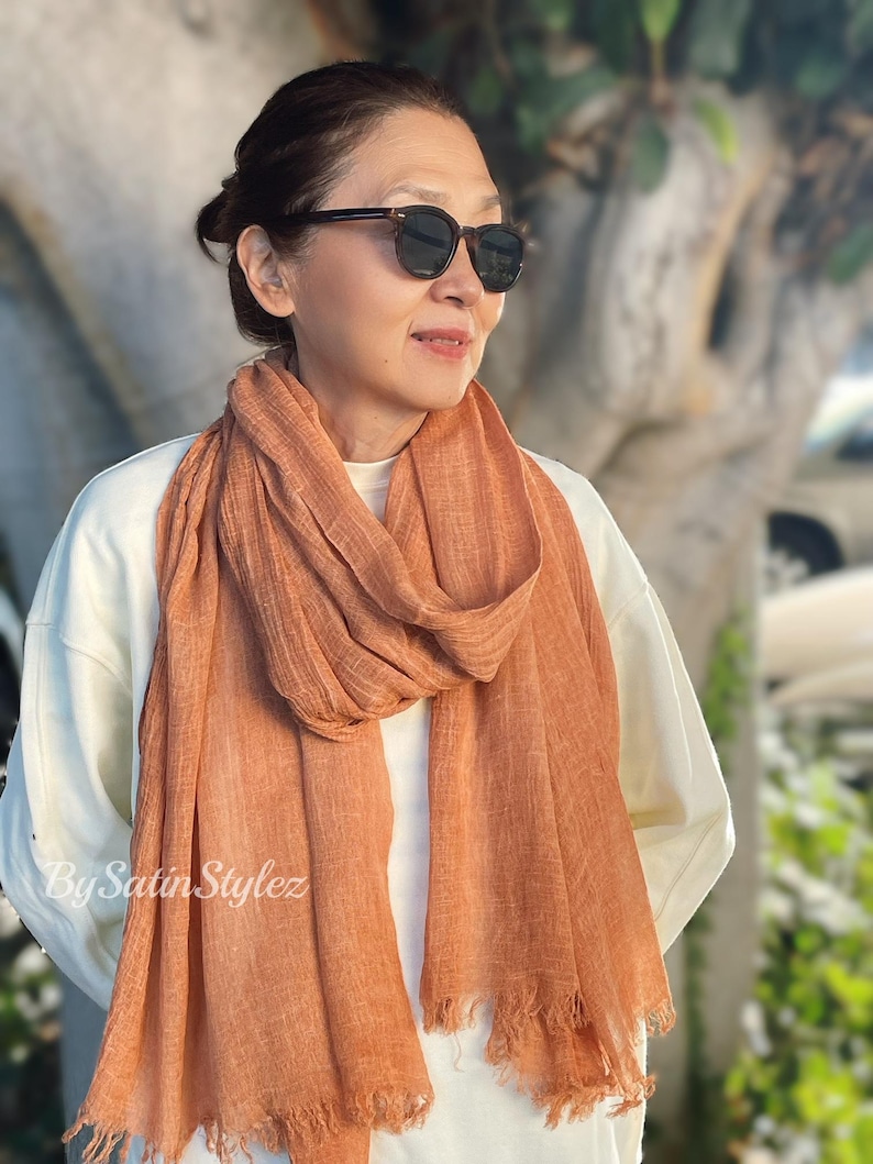 May include: A rust-coloured, fringed scarf made of a soft, textured fabric. The scarf is draped around the neck of a person wearing a white shirt and sunglasses.
