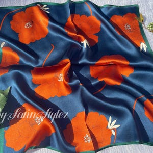 May include: A square silk scarf with a navy blue background and large orange floral patterns. The scarf has a green border and the text "By Satin Styler" in the lower left corner. The scarf is approximately 24 inches by 24 inches.