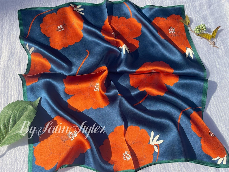 May include: A square silk scarf with a navy blue background and large orange floral patterns. The scarf has a green border and the text "By Satin Styler" in the lower left corner. The scarf is approximately 24 inches by 24 inches.