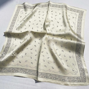 May include: A white silk bandana with a paisley pattern and a black border. The bandana is folded and laid flat on a white surface.