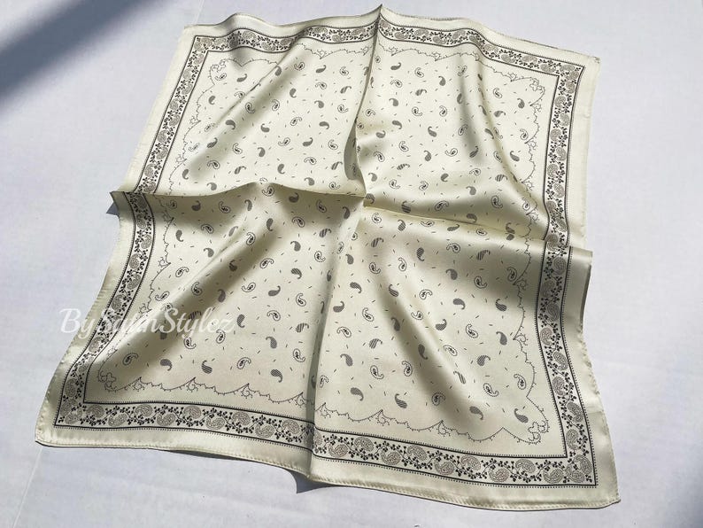 May include: A white silk bandana with a paisley pattern and a black border. The bandana is folded and laid flat on a white surface.