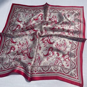 May include: A red and white paisley patterned silk scarf with a red border. The scarf is folded and laid flat on a white surface.