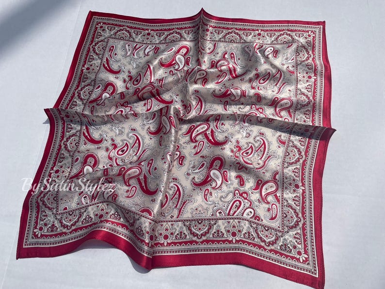 May include: A red and white paisley patterned silk scarf with a red border. The scarf is folded and laid flat on a white surface.