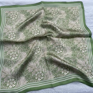 May include: A square, green silk scarf with a floral pattern. The scarf features a repeating design of flowers and foliage in shades of cream and green. The border is a darker green with white stripes.