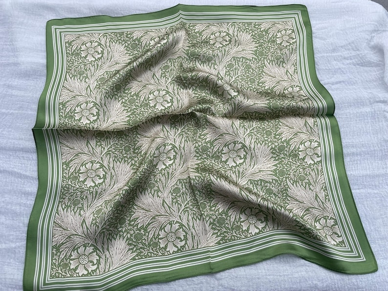 May include: A square, green silk scarf with a floral pattern. The scarf features a repeating design of flowers and foliage in shades of cream and green. The border is a darker green with white stripes.