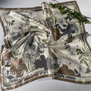 May include: Cream-colored square silk scarf with a floral and zebra print design. The scarf features brown, gray, and green floral patterns with a brown border. A sprig of greenery rests on the scarf.