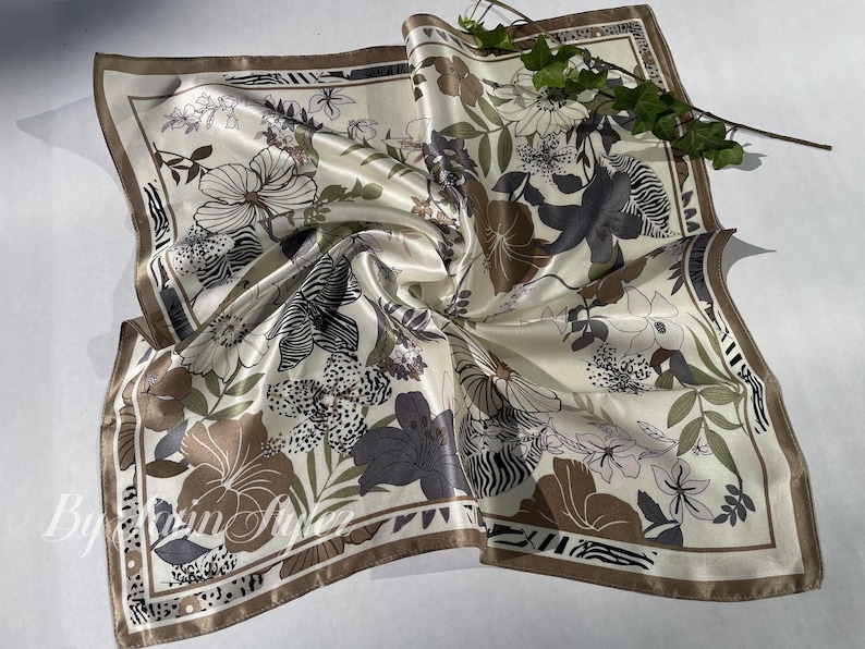 May include: Cream-colored square silk scarf with a floral and zebra print design. The scarf features brown, gray, and green floral patterns with a brown border. A sprig of greenery rests on the scarf.