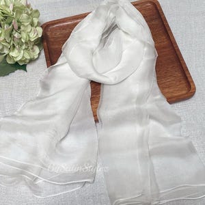 May include: A white silk scarf with a sheer, layered texture. The scarf is folded in half and draped over a wooden tray.