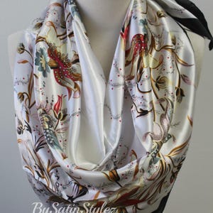 May include: White satin scarf with a floral print in shades of red, brown, green, and gold. The scarf has a black trim and the text "By Satin Stylez" is printed on the bottom.