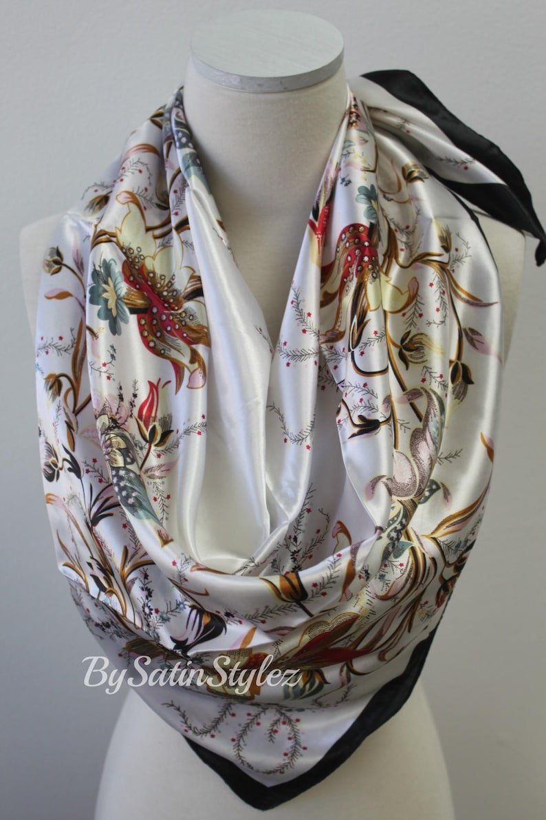 May include: White satin scarf with a floral print in shades of red, brown, green, and gold. The scarf has a black trim and the text "By Satin Stylez" is printed on the bottom.