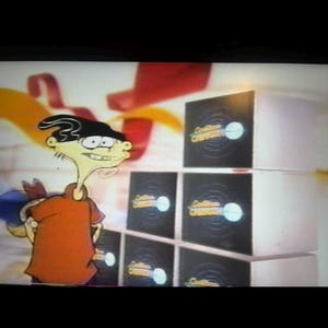 Peut inclure: Le personnage de dessin animé Ed d'Ed, Edd n Eddy se tient devant une pile de six boîtes noires et blanches. Chaque boîte porte un logo avec un cercle blanc et du texte. Ed porte une chemise orange.