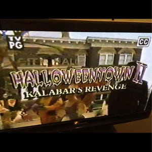 Puede incluir: Una captura de pantalla de la película "Halloweentown II: Kalabar's Revenge" en una pantalla de televisión. El título está en una fuente púrpura y naranja. La pantalla del televisor tiene una clasificación PG y el símbolo CC.